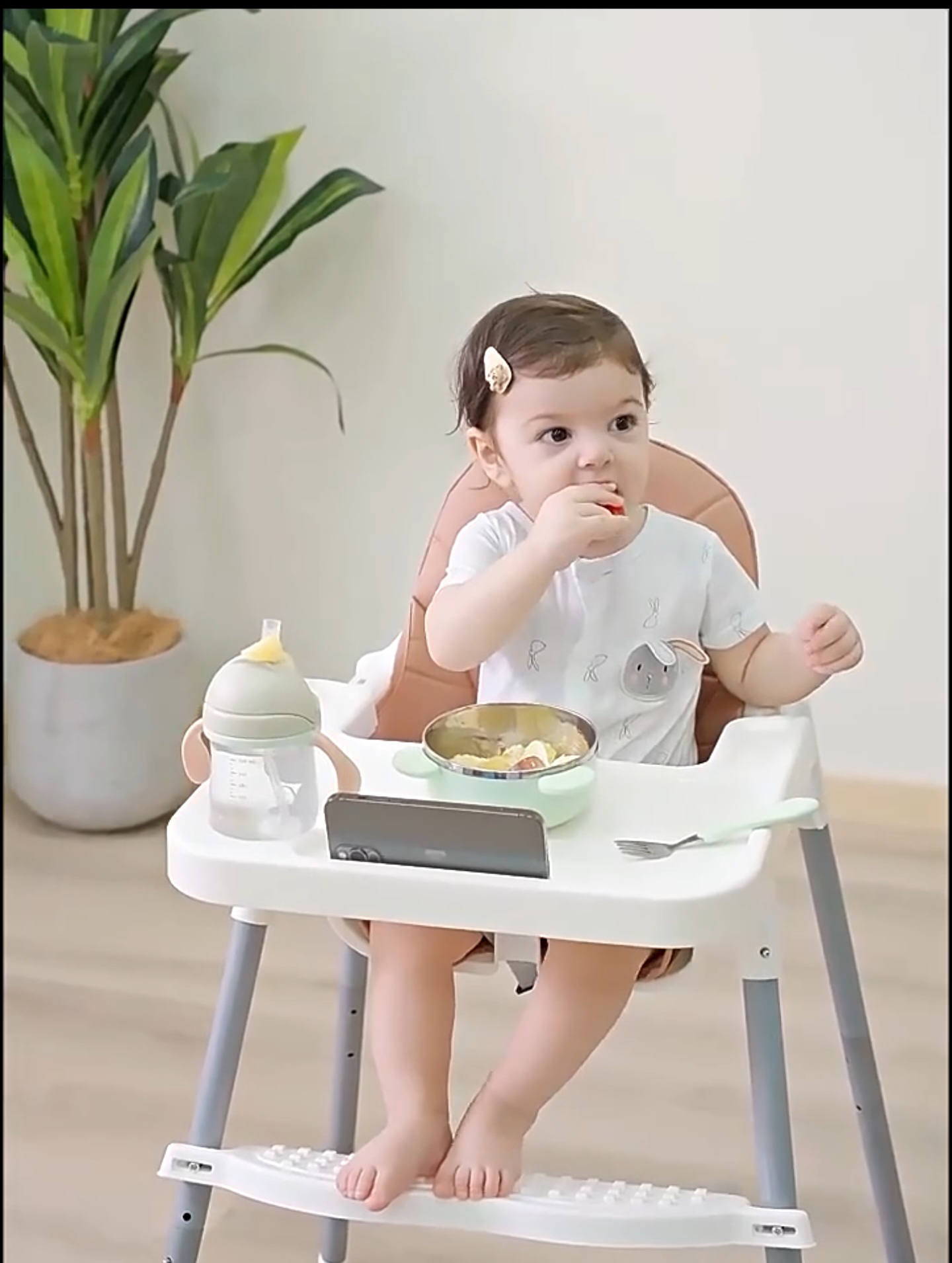 کودک در حال میل غذا با صندلی غذای کودک با سینی دارای جالیوانی و جای موبایا